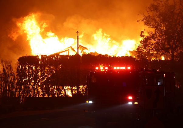 Fire truck with lights flashing parked before a large structure fire at night, flames and smoke filling the sky.