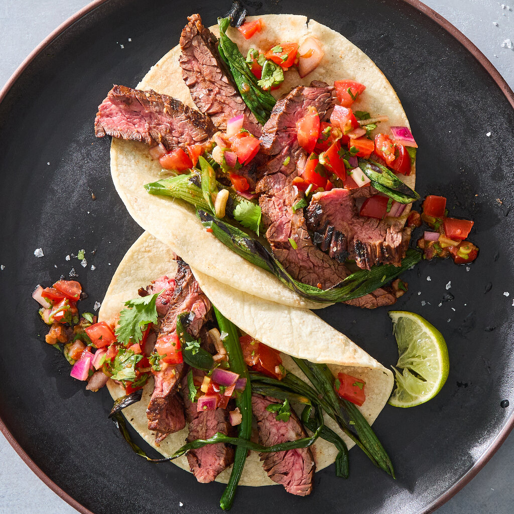 Two grilled steak tacos topped with charred green onions and tomato-onion salsa sit on a dark plate next to a lime wedge.
