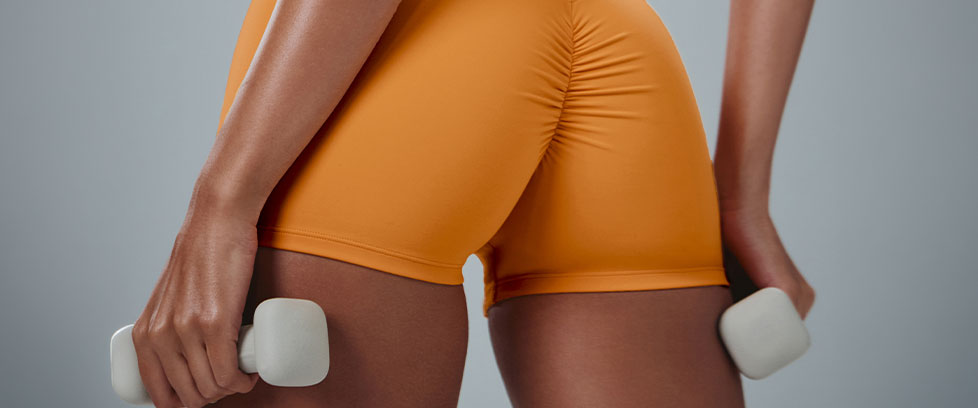 Person in orange scrunch butt biker shorts holds white dumbbells for a fitness strength workout.