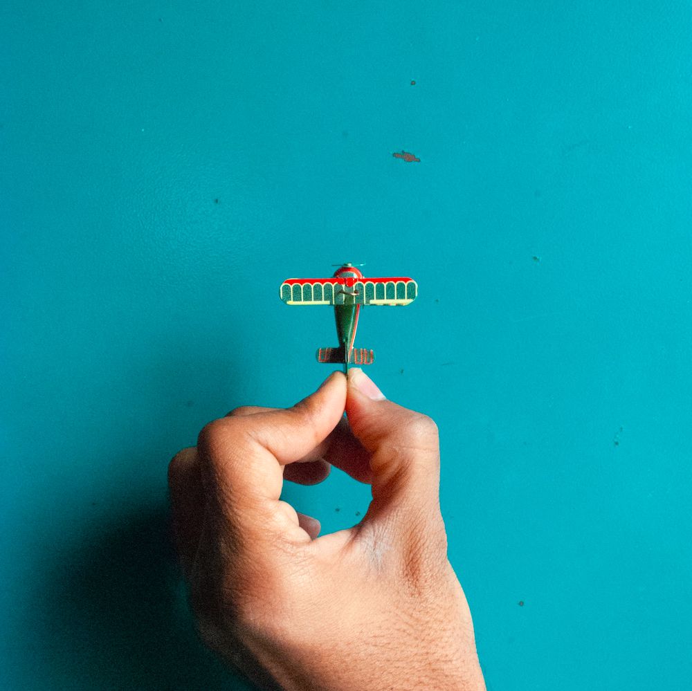 A hand holds a tiny green and red toy airplane against a teal background.