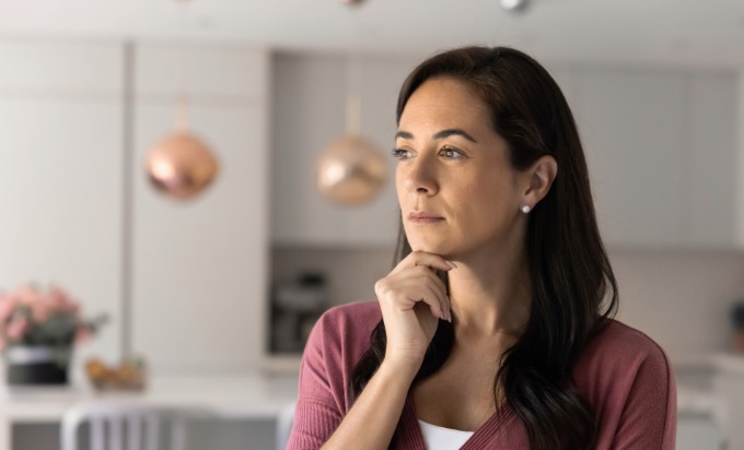 A pensive woman in a kitchen looks to the left, out a window, with her right hand on her chin  