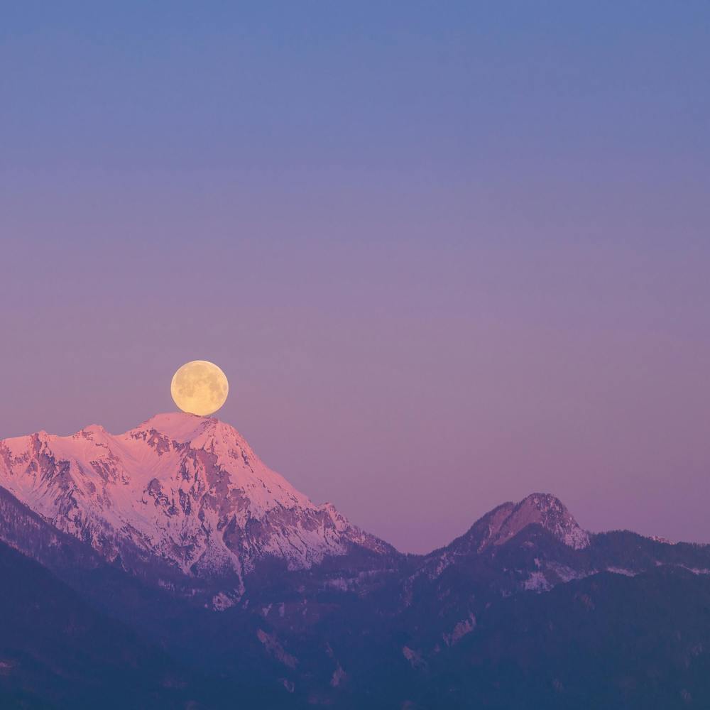The full moon appears to balance on the peak of a mountain, which looks pink in the sunset. 