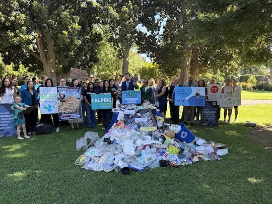 A group of policymakers and organization representatives stand in a park behind a large pile of single-use plastic waste holding signs in support of senate bill 54
