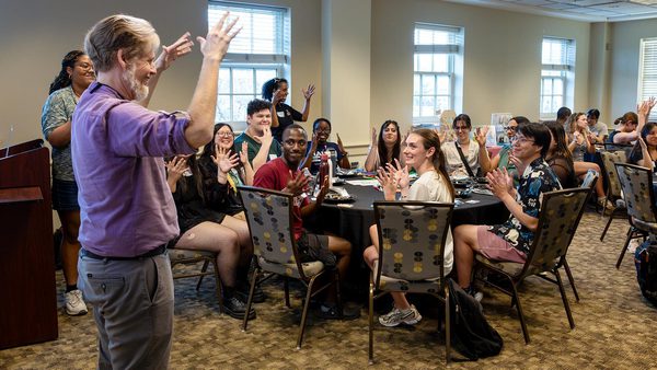 man in purple shirt signs at front of room as students at tables sign back