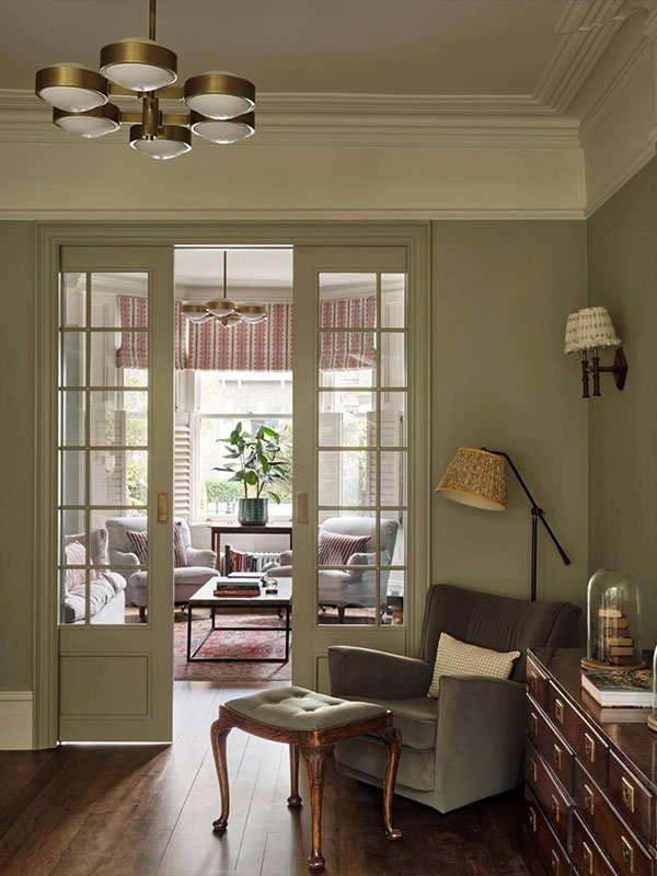 Two adjoining rooms with muted green walls, glass‑panelled doors, and vintage furniture. Brass lighting and soft upholstery create a classic, homely atmosphere.