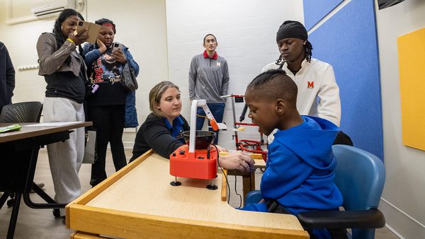 group helps young boy use 3D-printed feeder to eat, while two people record on smartphones