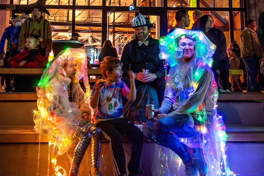 A family on the back deck of the Monterey Bay Aquarium dressed up in costumes lit up with colorful string lights on Halloween Member's Night