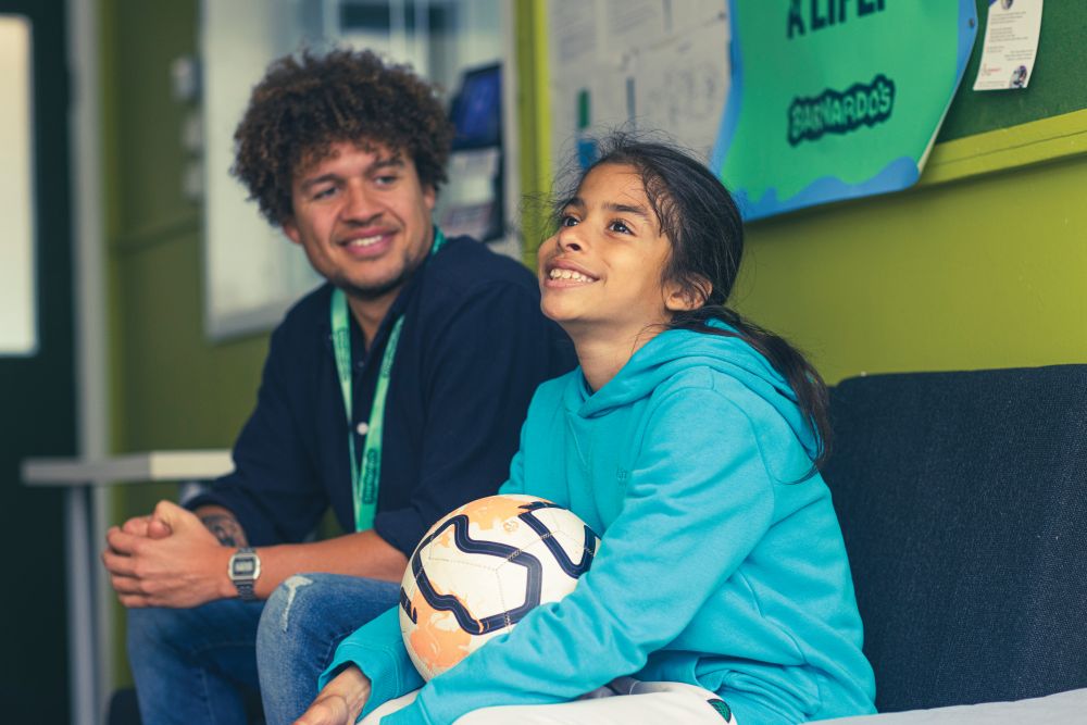 A young girl smiling holidng a football, sat next to a Barnardo's support worker