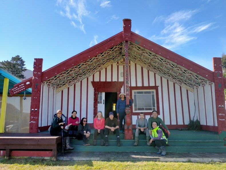 People sitting on the step in front of a wharenui (meeting house).
