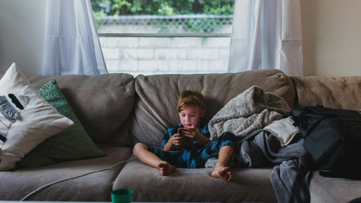 A young child sits in the middle of a large, puffy gray couch, staring intently into a phone.