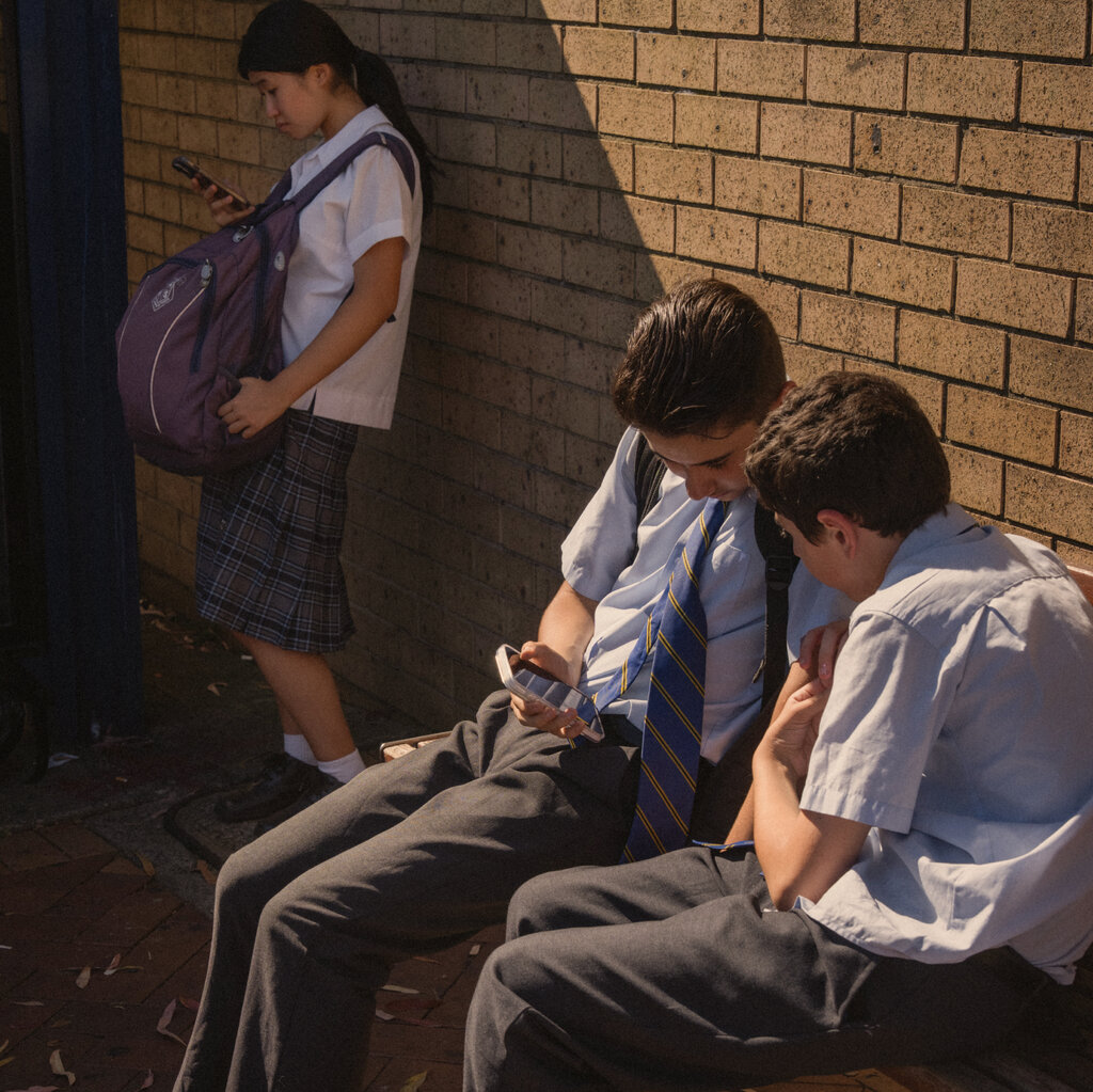 Two schoolboys in uniforms are sitting on a bench and looking at their cellphones. A schoolgirl standing nearby is doing the same.