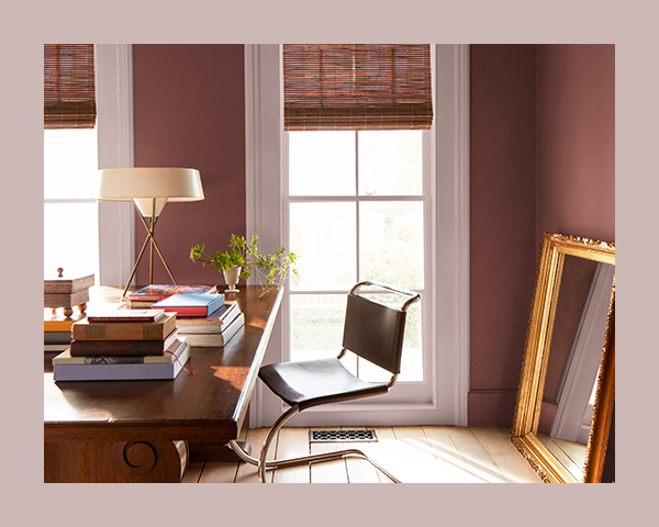 A reddish-brown home office with a wood desk topped with piles of books, a plant, and a lamp with a modern circular lampshade.