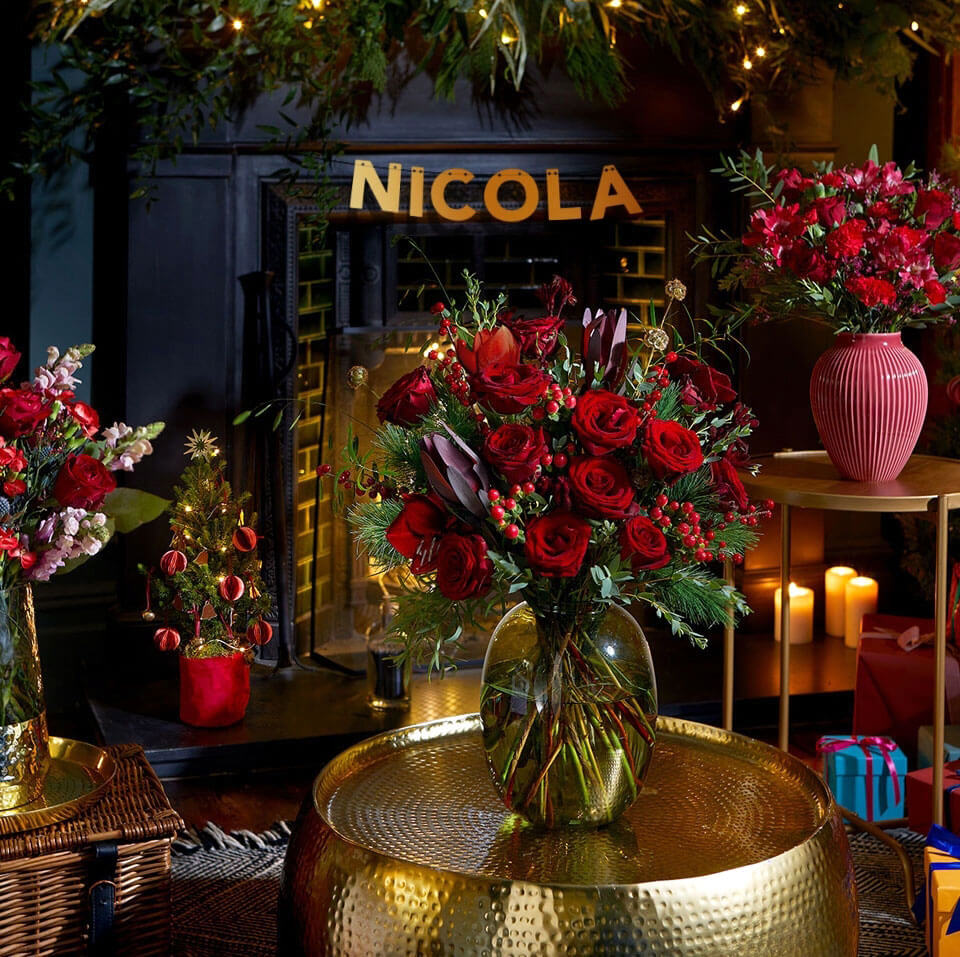 A festive bouquet of red roses in a clear vase, placed on a golden coffee table in front of a fireplace decorated with a garland and your name.