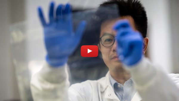 researcher in white coat and blue gloves holds up bioplastic, with red play button