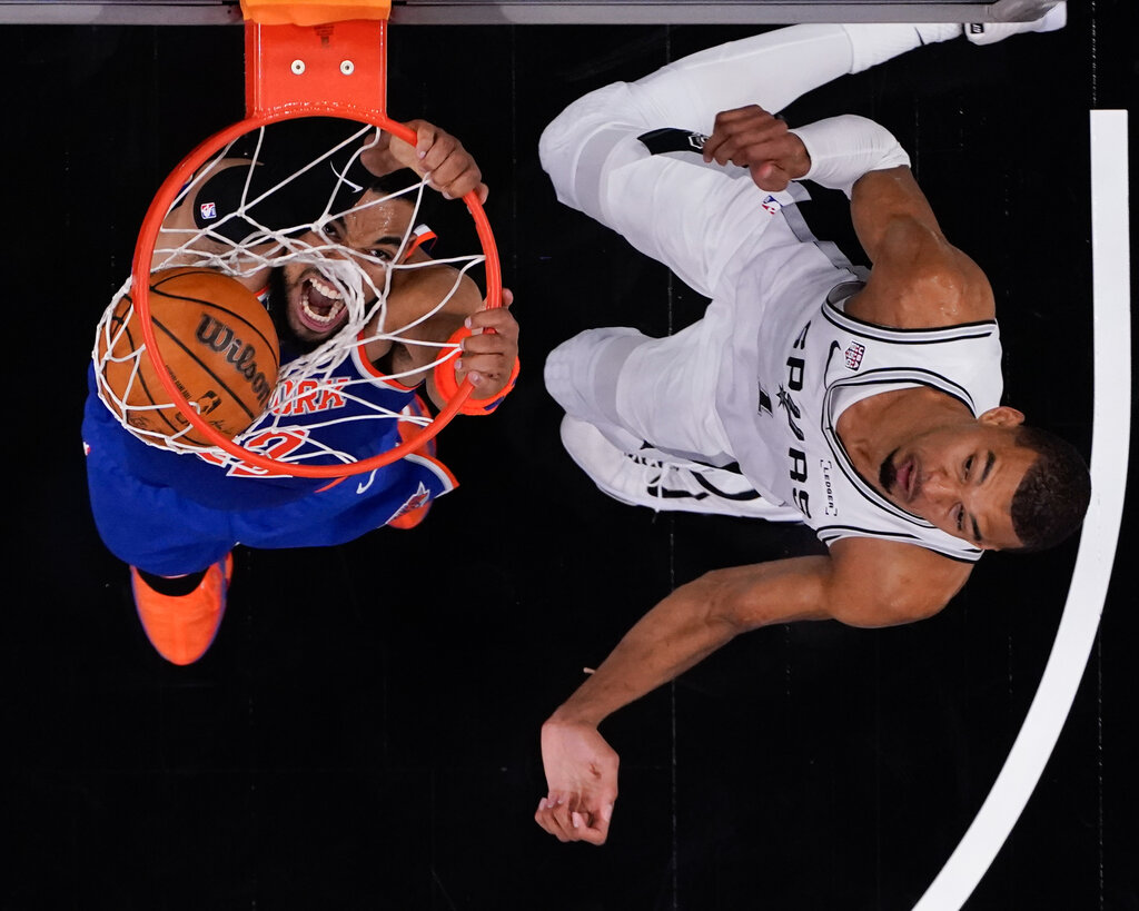 Overhead view of a person in a blue and orange uniform hanging from a basketball hoop with a ball. A person in white looks up from the court.