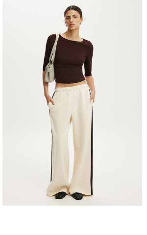 Woman wearing a brown cropped long-sleeve top and cream wide-leg trousers with dark side stripe, holding a beige shoulder bag