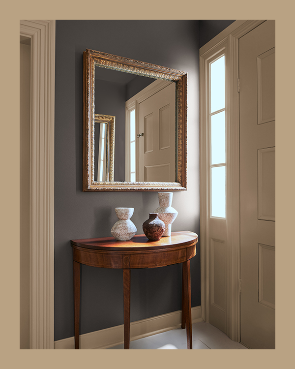 An entryway with brown painted walls, a half-moon wood accent table, and large mirror framed in gold.