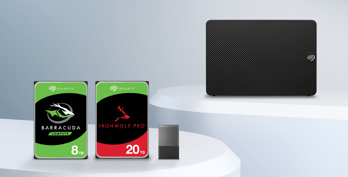 Two Seagate hard drives, a Barracuda 24TB and IronWolf Pro 20TB, sit on a white surface beside a compact black external drive, all against a soft gray background.