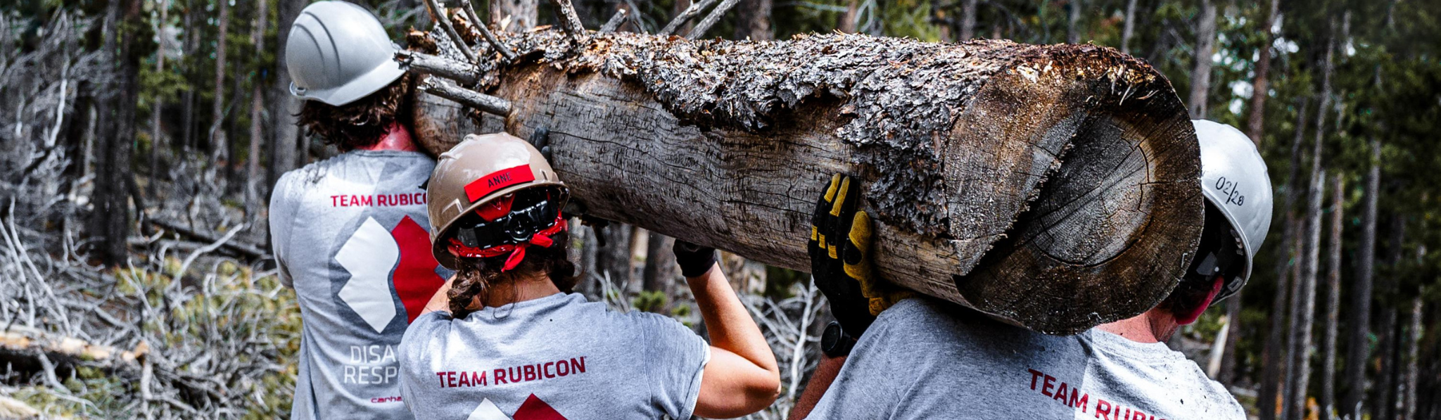 Three Greyshirts carry a massive piece of burned wood. The wood is horizontal and they're carrying it on their shoulders. They are all wearing grey shirts and hard hats.