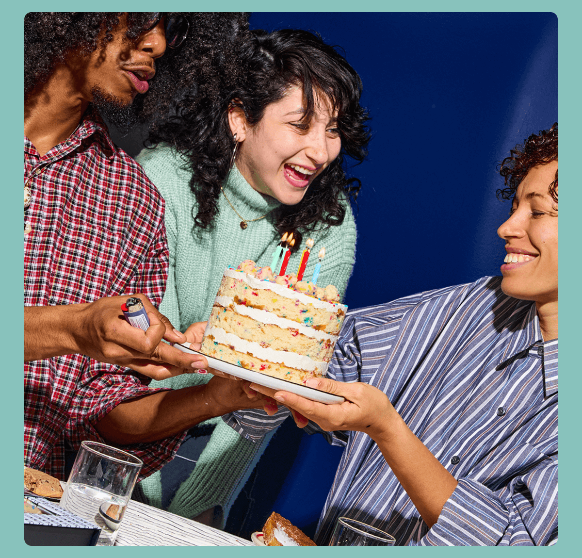 Three people gathered around a table celebrate with a layered Birthday Cake topped with lit candles. One person holds the cake on a plate while another holds a lighter near the candles, and a third smiles toward the cake. Glasses and dessert plates sit on the table against a blue background.