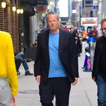 Matthew Perry, wearing dark pants, a dark blazer and a light blue T-shirt, walks on a crowded city sidewalk.
