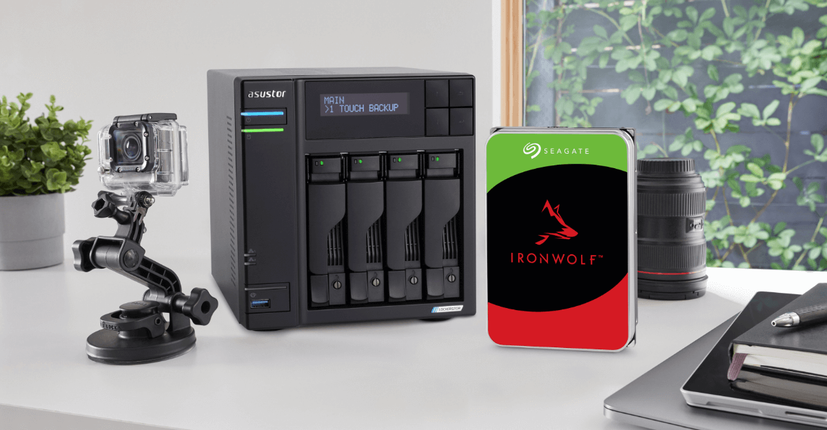 A desktop setup featuring an Asustor NAS enclosure, a Seagate IronWolf hard drive, a mounted action camera, a camera lens, and notebooks.