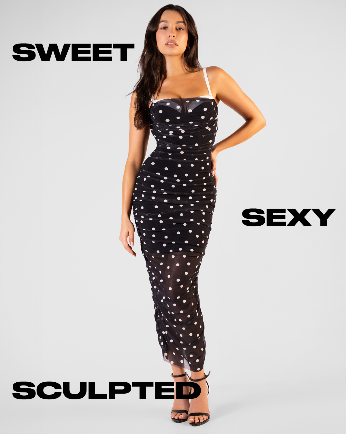 A model wearing a black polka dot dress with a fitted silhouette and strappy heels.