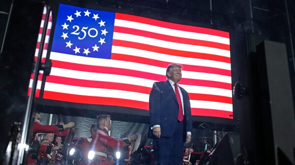 President Trump stands on a stage with a flag resembling the American flag but where the stars are it says “250” with stars in a circle around that number.