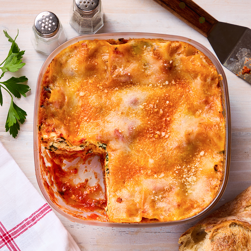 A golden, bubbly lasagna cooked in the microwave inside a square Anyday dish, with one slice removed to reveal layers of pasta, sauce, cheese, and greens. The dish sits on a light wood table next to bread, parsley, and a red-striped napkin.