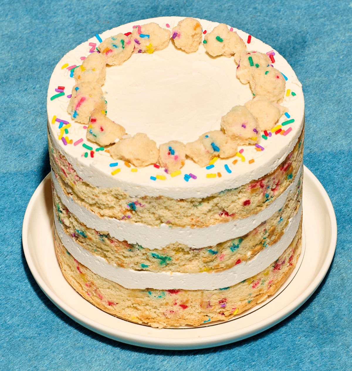 Close-up of a layered birthday cake with white frosting and rainbow sprinkles on a white plate against a blue background.
