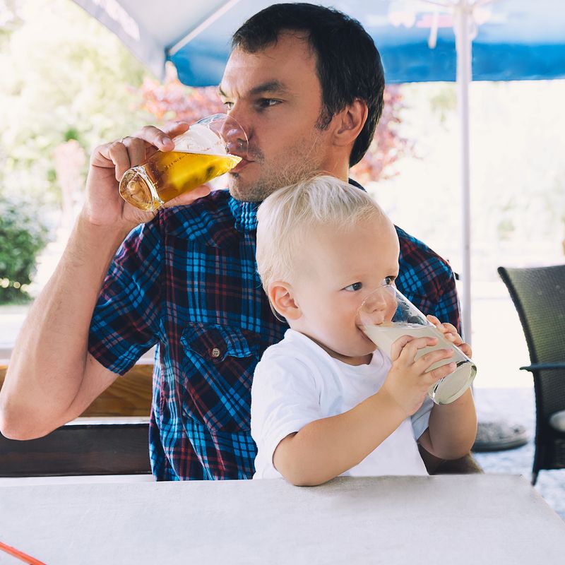 An adult drinking a beer with a toddler drinking a juice on his lap.
