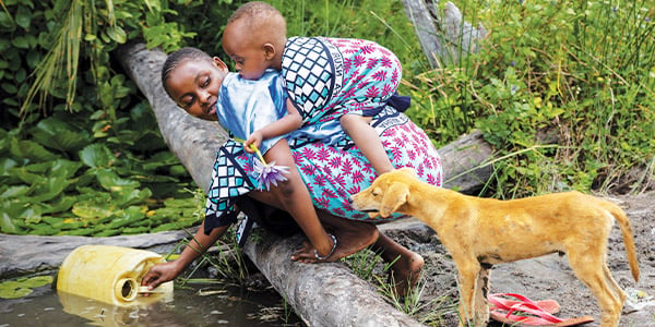 A mother leans over to collect dirty water from a pond while carrying her son on her back. 
