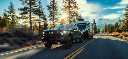 A Chevy truck pulls a trailer through a scenic woodland setting with mountains rising in the distance.