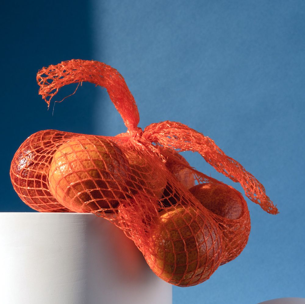 Oranges in a red mesh bag sit on top of a white block in front of a dark blue background.
