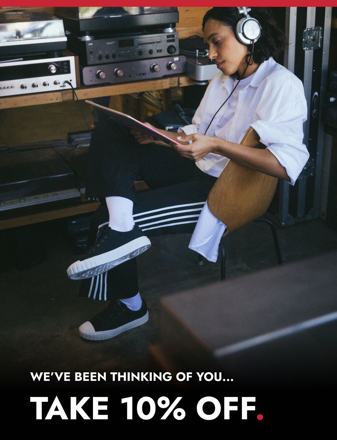 A person wearing black Red Ball Jets sneakers sits in a wooden chair with headphones on, holding a vinyl record while surrounded by audio equipment. Text at the bottom reads, “WE’VE BEEN THINKING OF YOU… TAKE 10% OFF.”