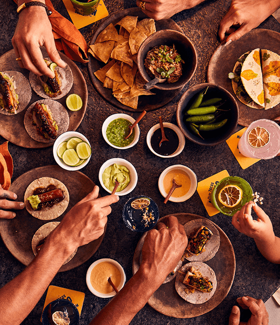 An inviting spread of tacos, dips, and drinks surrounded by hands reaching in to share the vibrant feast.