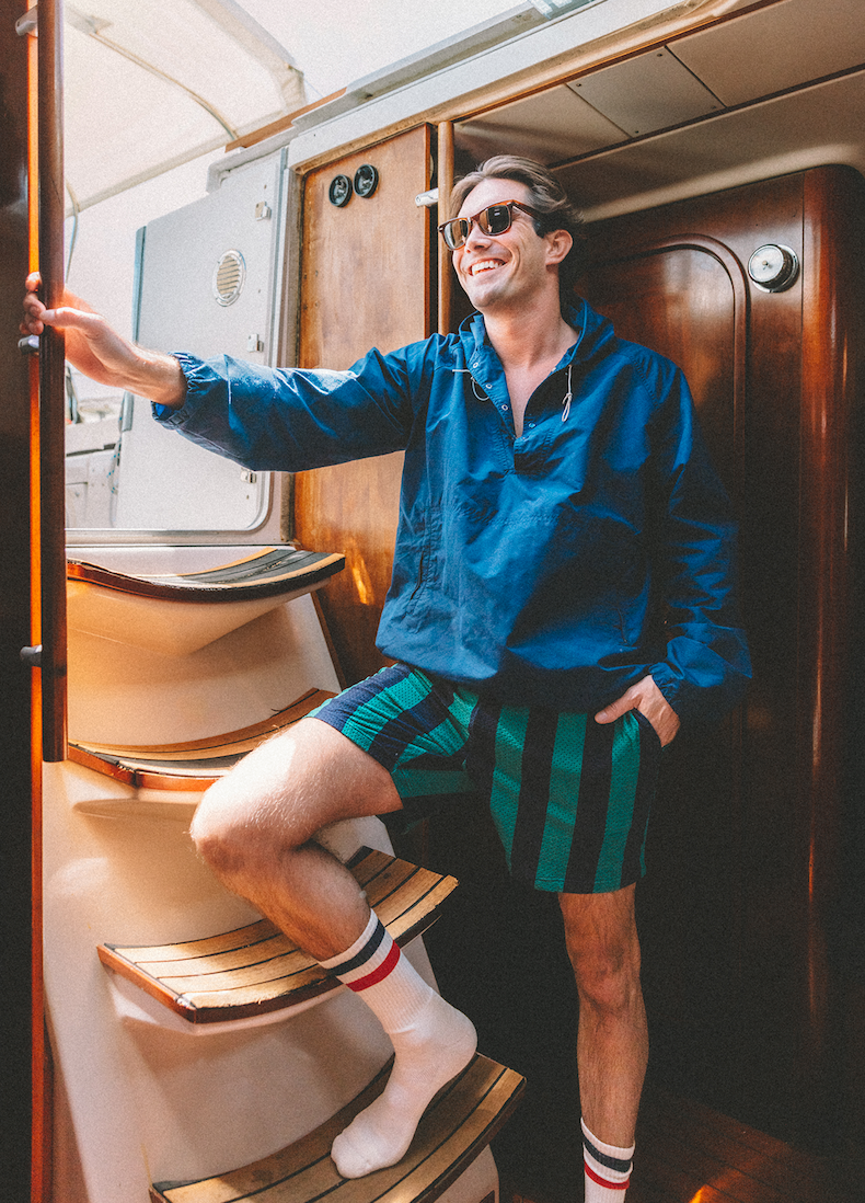 Man inside boat wearing sunglasses, blue jacket and green/navy stripe lounge shorts