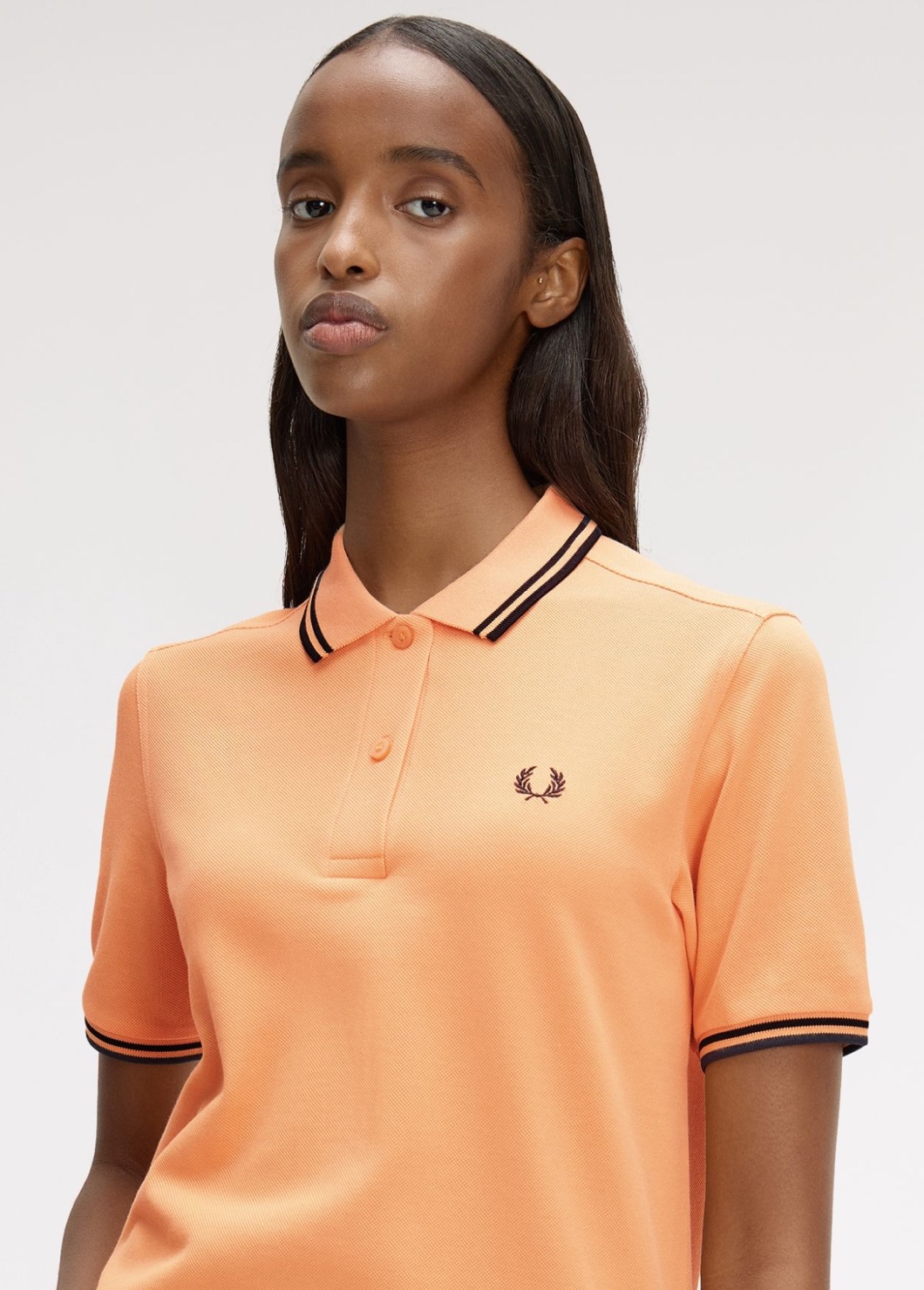 A female model wearing a Fred Perry Shirt in a vibrant peach colour with black twin tipping on the collar