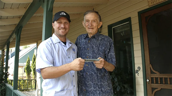 A young man and an older man hold a postcard while standing on a porch.