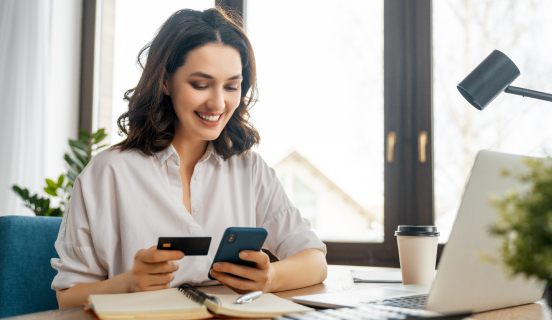 A smiling woman holds a credit card in one hand and her smartphone in the other, while she sits at an office desk.