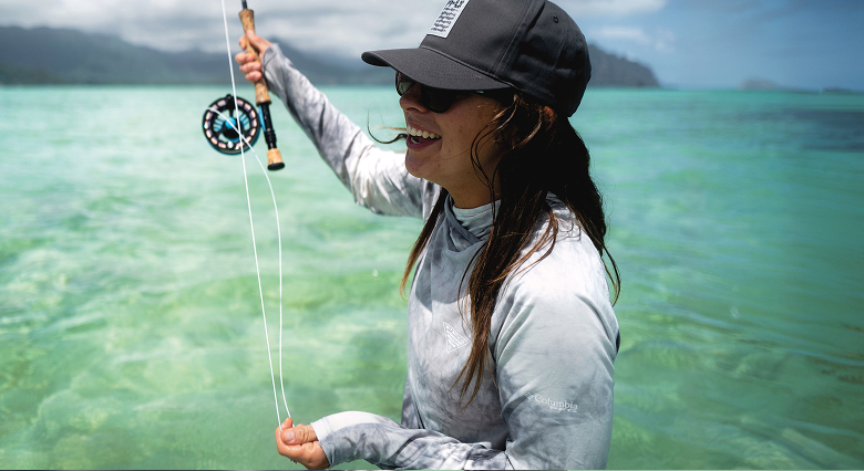 A woman standing in the water, wearing a light blue sun shirt, black ball cap, and sunglasses, holding a fishing rod.