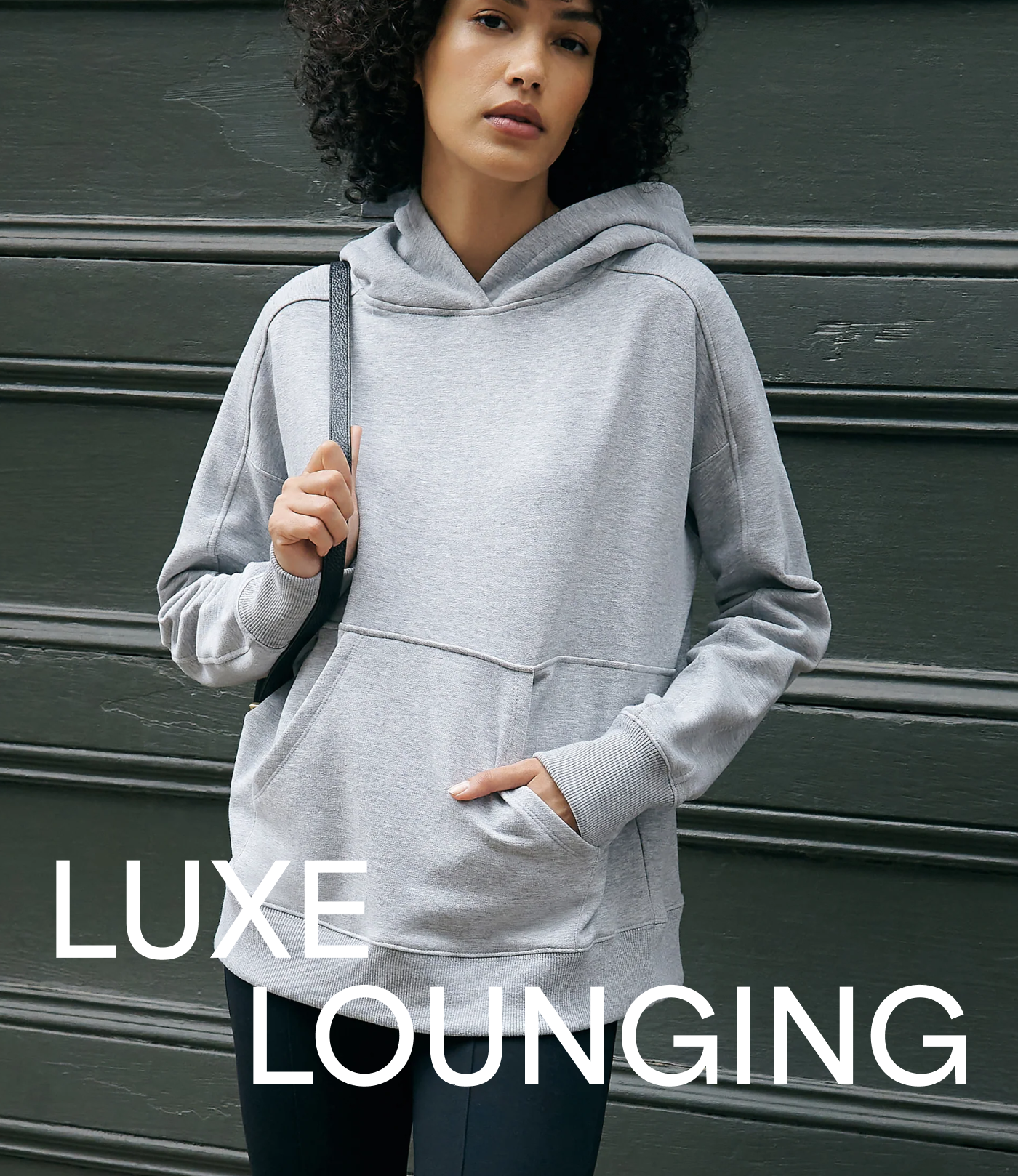 Astrid in The Ida Hoodie with a kangaroo pocket stands against a dark paneled wall with one hand in her pocket, with large overlaid text reading “Luxe Lounging.”