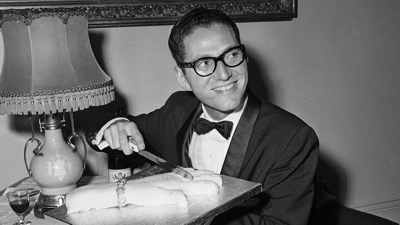 Tom Lehrer cutting a cake in the shape of a woman's hand while backstage at the Palace Theatre, London, 1959
