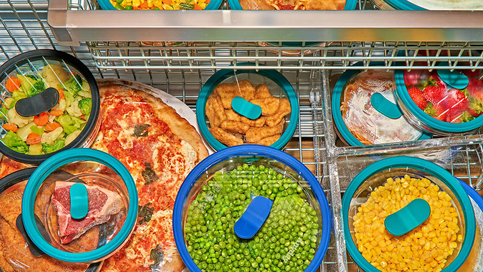 A packed freezer filled with meals and ingredients stored in plastic-free Anyday Everything Dishes. Glass containers hold frozen peas, corn, strawberries, cooked pasta, and breaded chicken, alongside a frozen pizza and raw steak, showcasing a sustainable and airtight food storage solution.