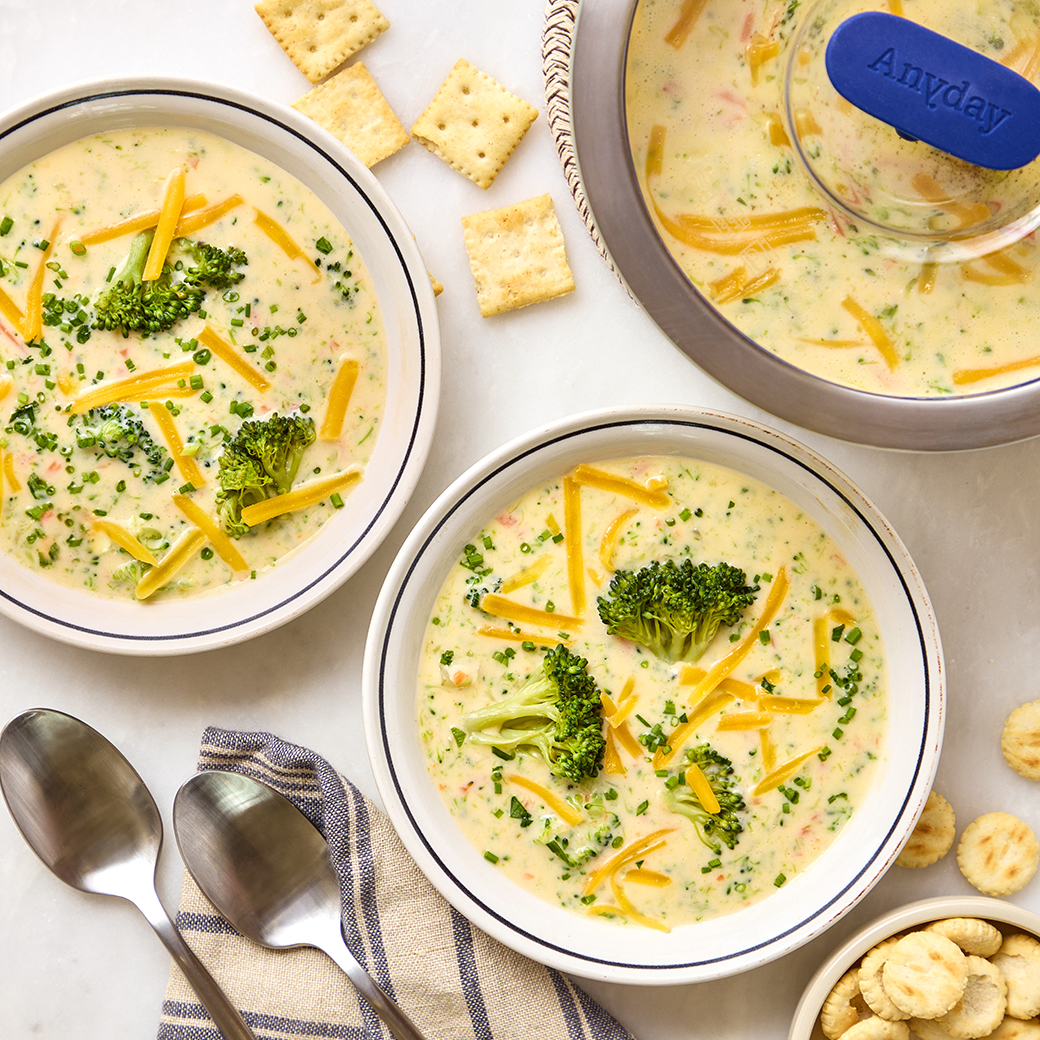 Creamy broccoli cheddar soup served in bowls alongside an Anyday Large Deep Dish with a blue glass lid; a plastic-free way to cook in the microwave and serve cozy meals.