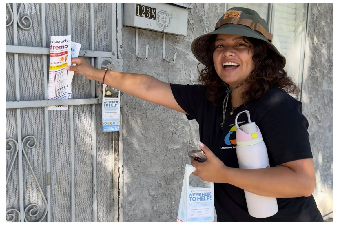 A community health worker wearing a bucket hat and holding a lavendar insulated water bottle puts a pamphlet behind a white residential screen door.