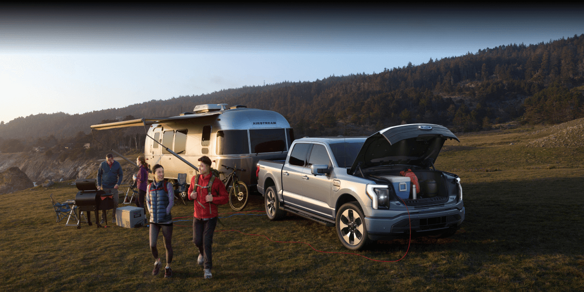 A group of people parked in a field, gray F-150 Lightning and Airstream camper behind them.