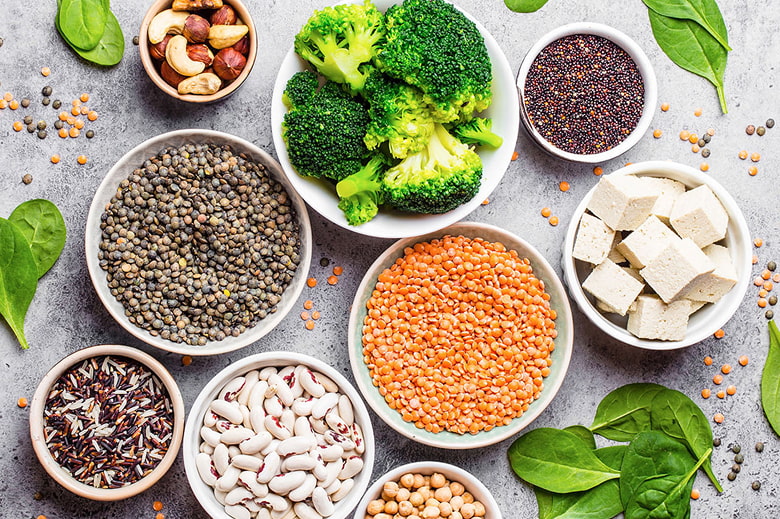 A top-down view of various bowls filled with colorful vegan ingredients, including green and red lentils, white beans, chickpeas, quinoa, wild rice, and broccoli, showcasing a variety of healthy meat alternatives.