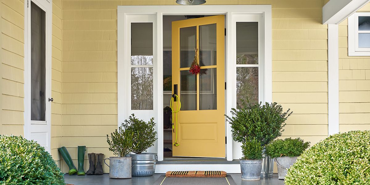 The front porch of a yellow-painted house with a yellow door, white trim, plants in metal pails, and two round shrubs out front.