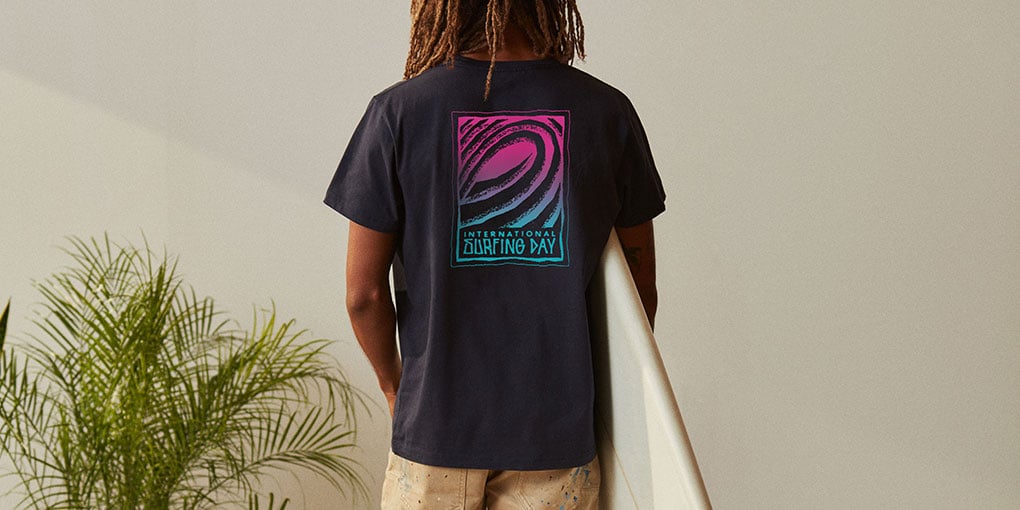 A model with long dreads holds a surfboard while wearing a black t-shirt with a surfrider logo on the back.